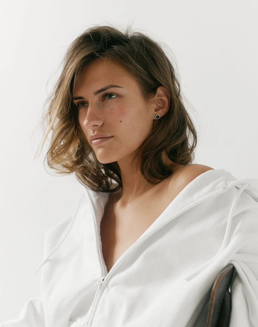 A woman with shoulder-length, tousled hair gazes thoughtfully off to the side, wearing a white hoodie. - Chemical Peels in New York, NY