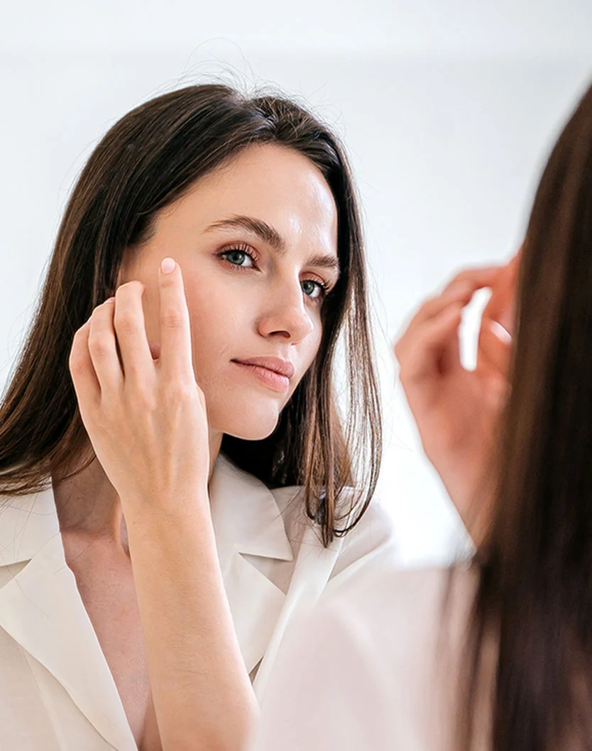 A woman with long brown hair examines her reflection in a mirror, gently touching her face with a thoughtful expression. - Eyebrow Transplantation in New York, NY