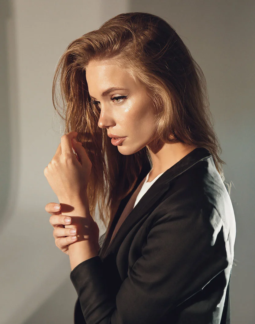 A woman with long, wavy hair in a black blazer gently touches her hair, illuminated by soft lighting. - Facial Filler in New York, NY