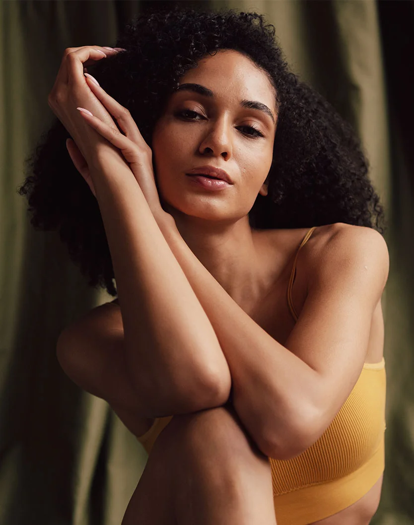 A woman with curly hair poses gracefully, resting her arms on her knees, wearing a yellow tank top against a soft backdrop. - Facial Liposculpture in New York, NY