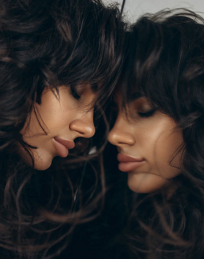 Two women with long, curly hair lean in close, their faces touching softly, showcasing their striking features and intimate connection. - Lip Reduction in New York, NY