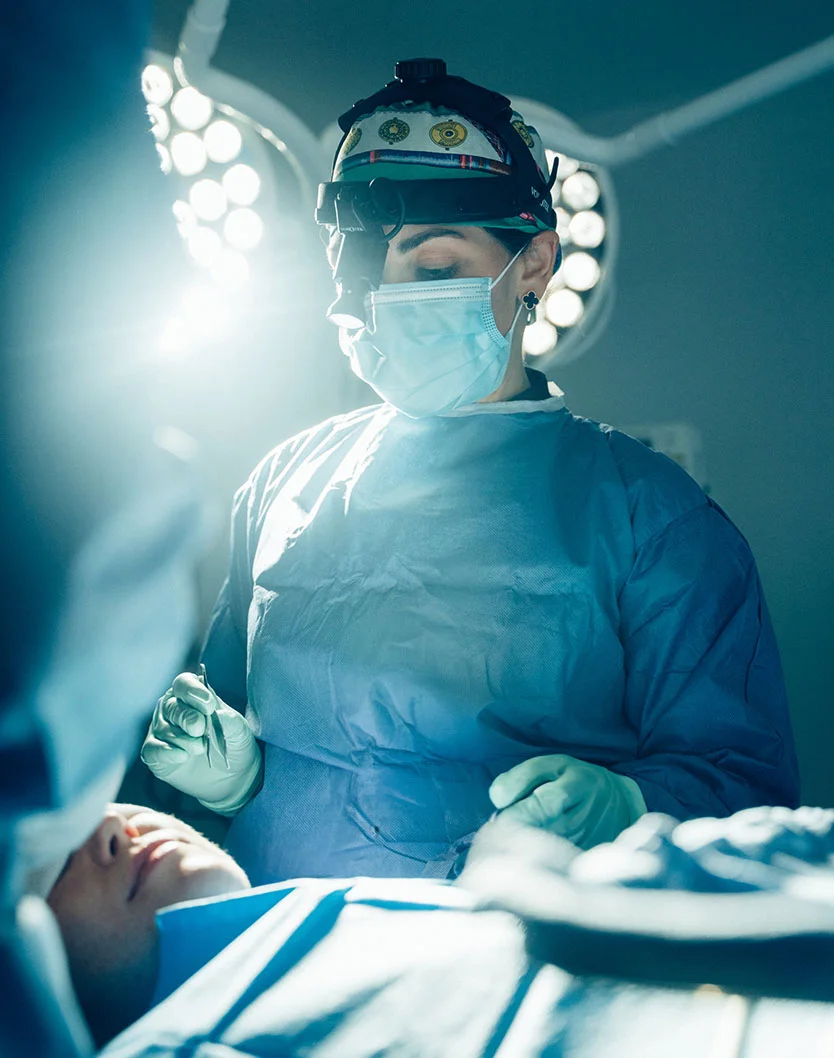 A medium shot of Dr. Rabach in a sterile surgical setting, wearing a light blue surgical gown, gloves, face mask, and cap. Overhead surgical lights highlight both the surgeon and patient. The foreground is blurred, focusing on the surgeon’s precise actions. The clean, organized background reflects the professionalism essential in surgical environments. - Revision Facelift in New York, NY