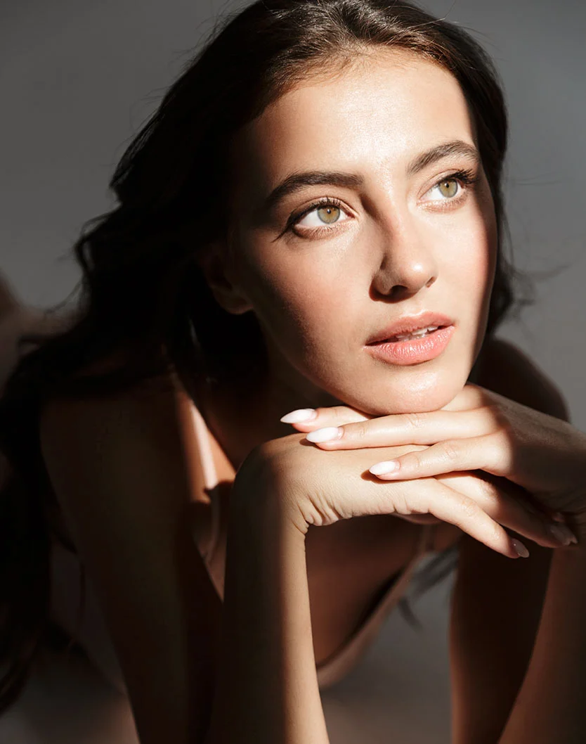 A close-up of a woman resting her chin on her hands. She has long, dark brown hair and light-toned skin, with golden-green eyes looking upward and to the right. Her hands rest neatly on her cheeks, with light nails. Dramatic lighting highlights her neck and shoulders, set against a neutral gray background, creating a graceful and tranquil mood. - Skar Removal in New York, NY