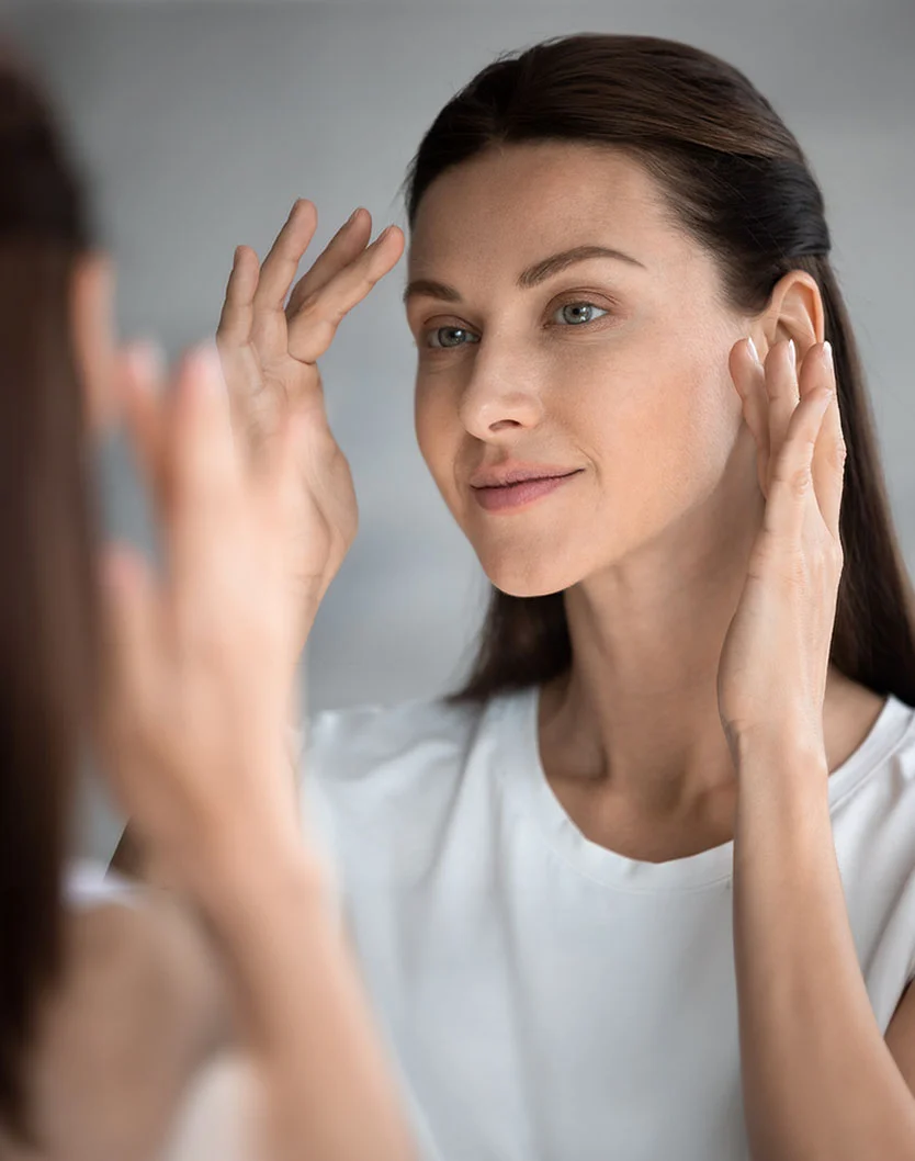 A medium close-up captures a woman with light brown auburn hair looking at her reflection in a mirror. She gently touches her forehead and cheeks with her hands, her expression calm and neutral. Wearing a light cream or white t-shirt, she stands in a softly lit bathroom-like setting with a light gray, blurred background. The partial reflection of her upper body and hands in the mirror emphasizes her focus on herself in the moment. - Skin Cancer Screening in New York, NY