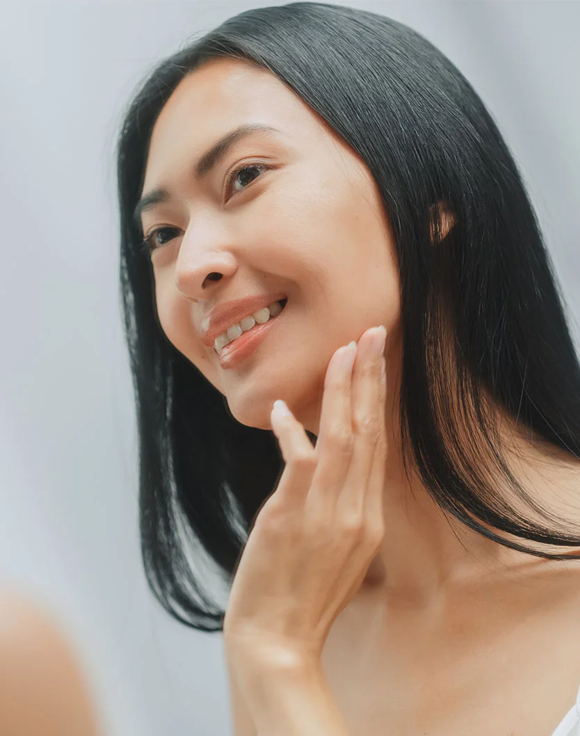 A close-up of a woman with dark shoulder-length hair and a light complexion examining her face in a mirror. She gently touches her neck and jawline with a slight smile. Soft, natural lighting highlights her features, while the blurred light gray or white background emphasizes the focus on self-care and personal grooming. - Ultherapy in New York, NY