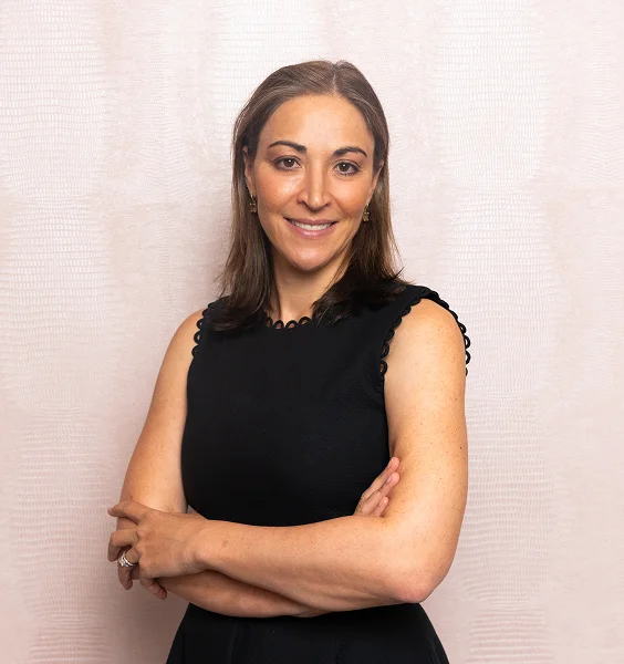 Professional woman in black sleeveless dress with arms crossed smiling against white brick wall