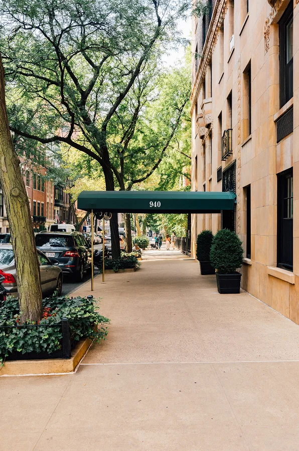 Urban sidewalk with covered entrance, trees, and parked cars between residential buildings