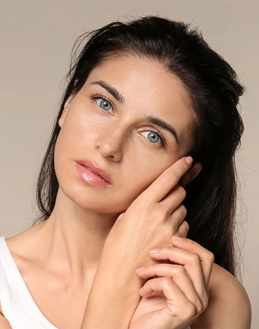 Woman with dark hair and blue eyes touching face, showcasing natural beauty and clear skin on neutral background