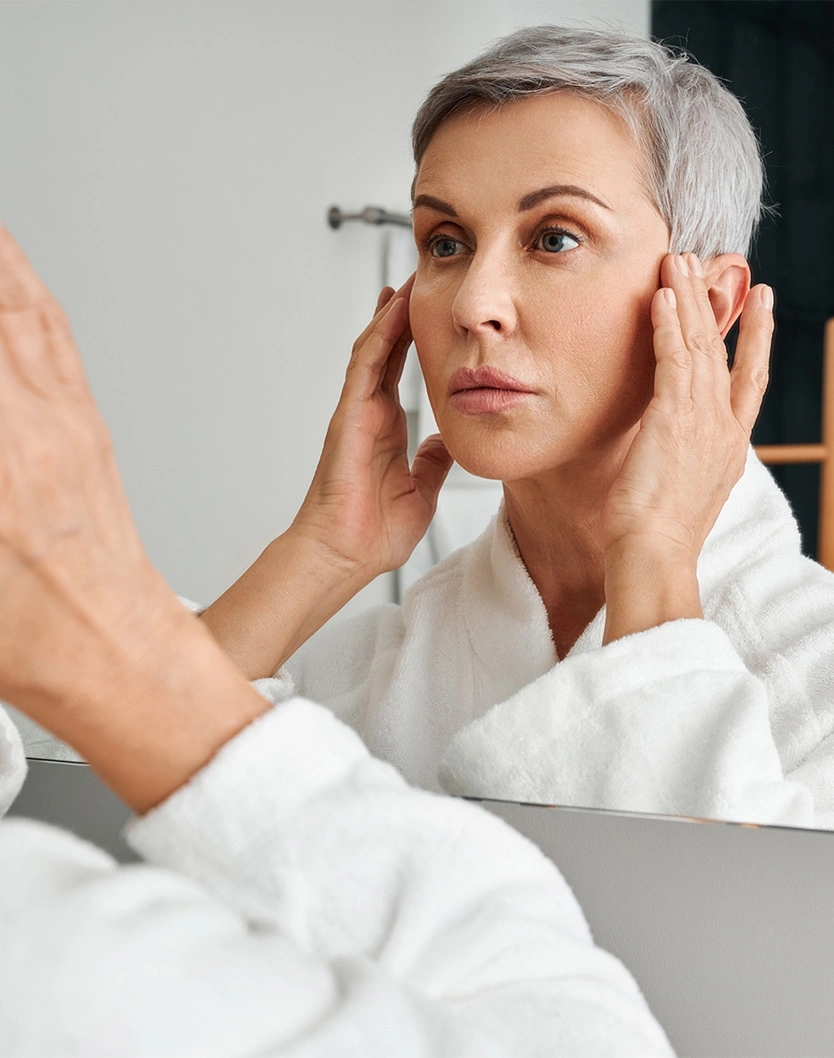 Woman with short platinum hair examining face in mirror while applying skincare treatment in white bathrobe