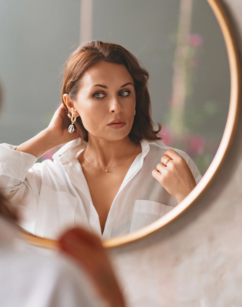 Woman in white shirt looking at reflection in round gold mirror while touching earring