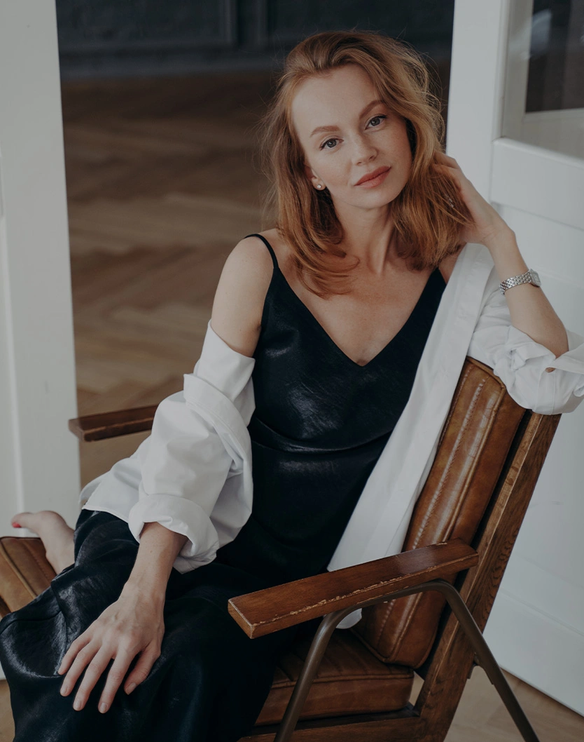 Red-haired woman in black dress and white cardigan sitting on wooden chair in natural indoor lighting