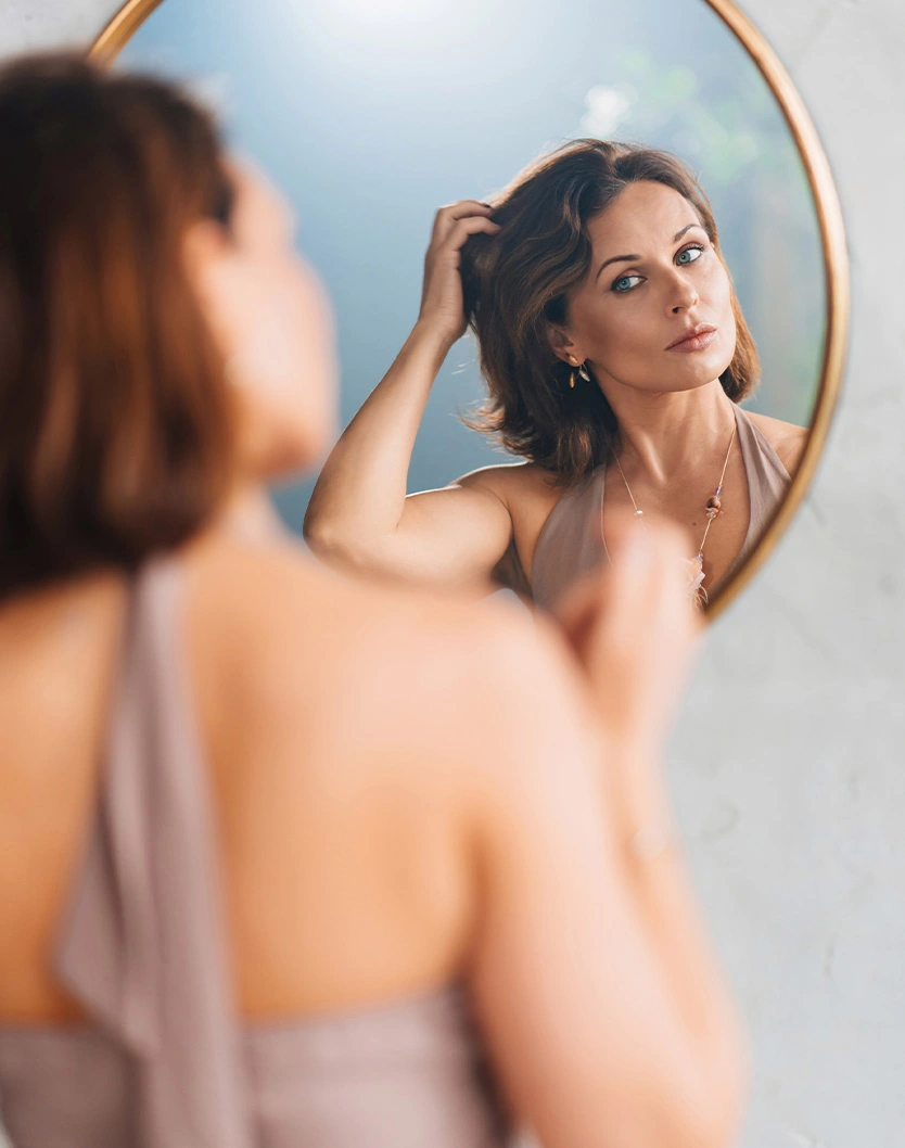 Woman examining her reflection in round mirror while touching her hair in natural light