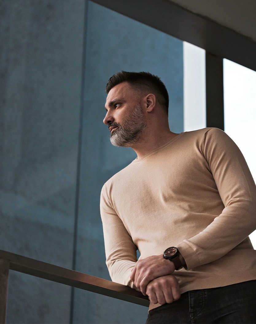 Man in beige sweater standing by railing looking away thoughtfully in modern interior with natural light