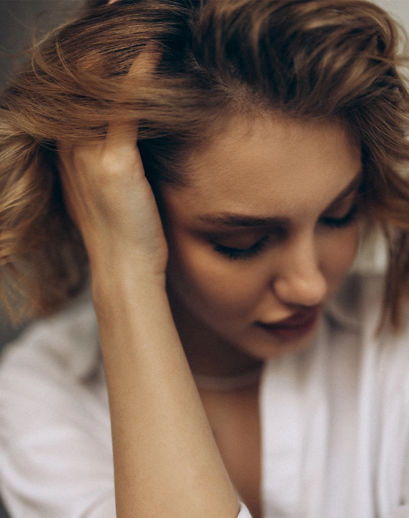 Woman with tousled brown hair looking down with hand in hair, appearing stressed or contemplative in white shirt