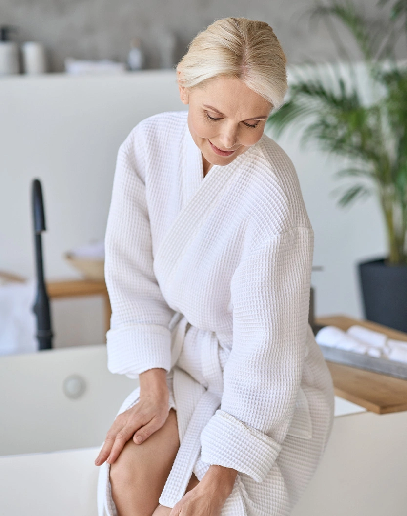 Woman in white waffle bathrobe sitting by bathtub in modern spa bathroom