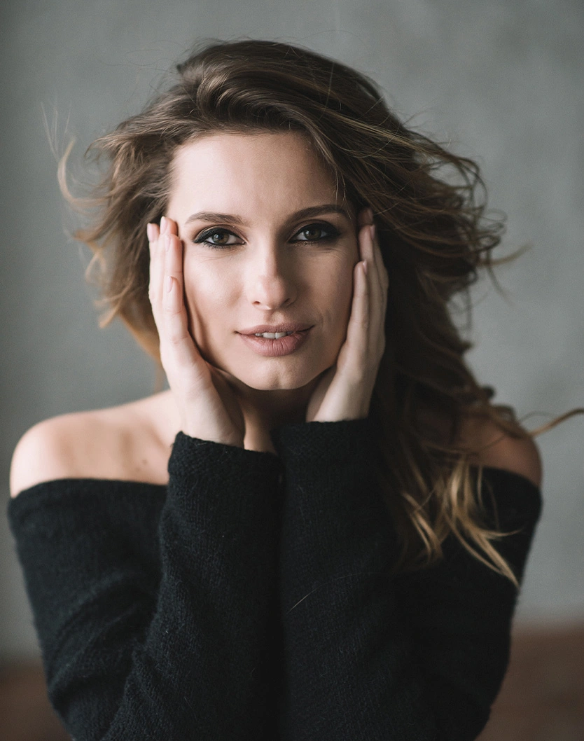 Woman with wavy brown hair wearing black off-shoulder sweater posing with hands near face against gray background
