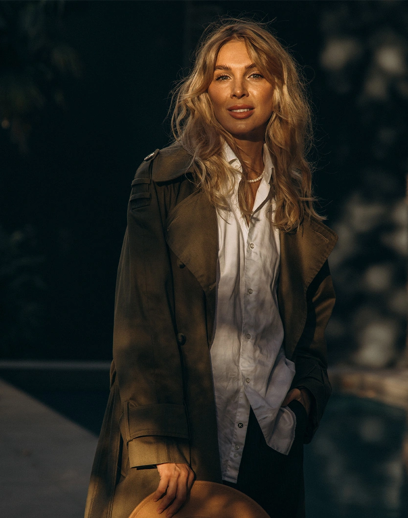 Woman in brown plaid blazer and white shirt with blonde wavy hair smiling outdoors in natural lighting