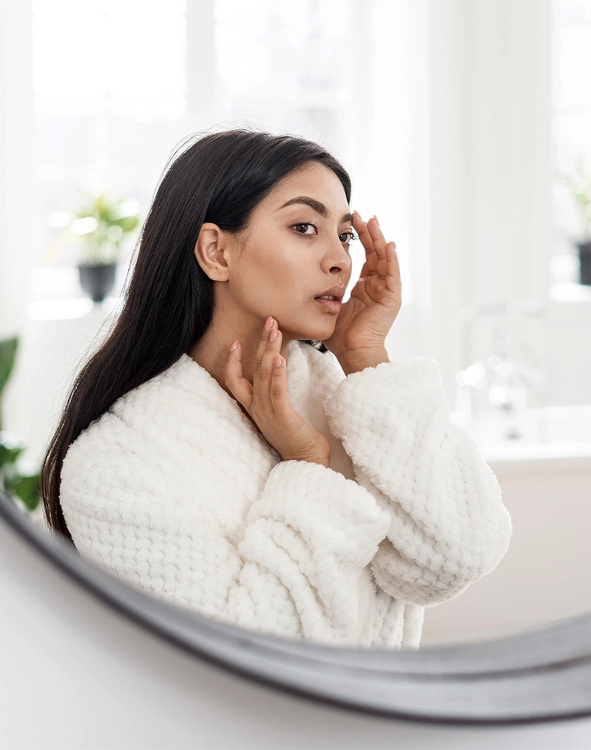 Woman in white bathrobe examining skin in bathroom mirror during skincare routine