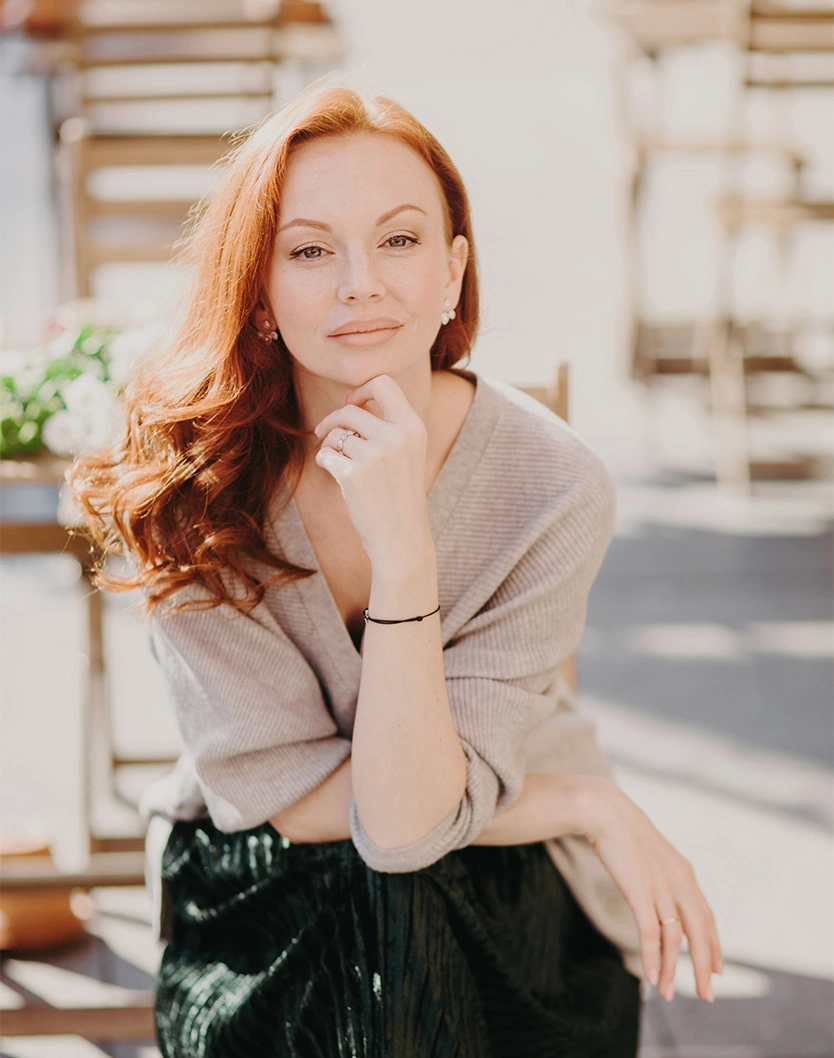 Red-haired woman in beige sweater sitting outdoors with hand on chin in natural lighting