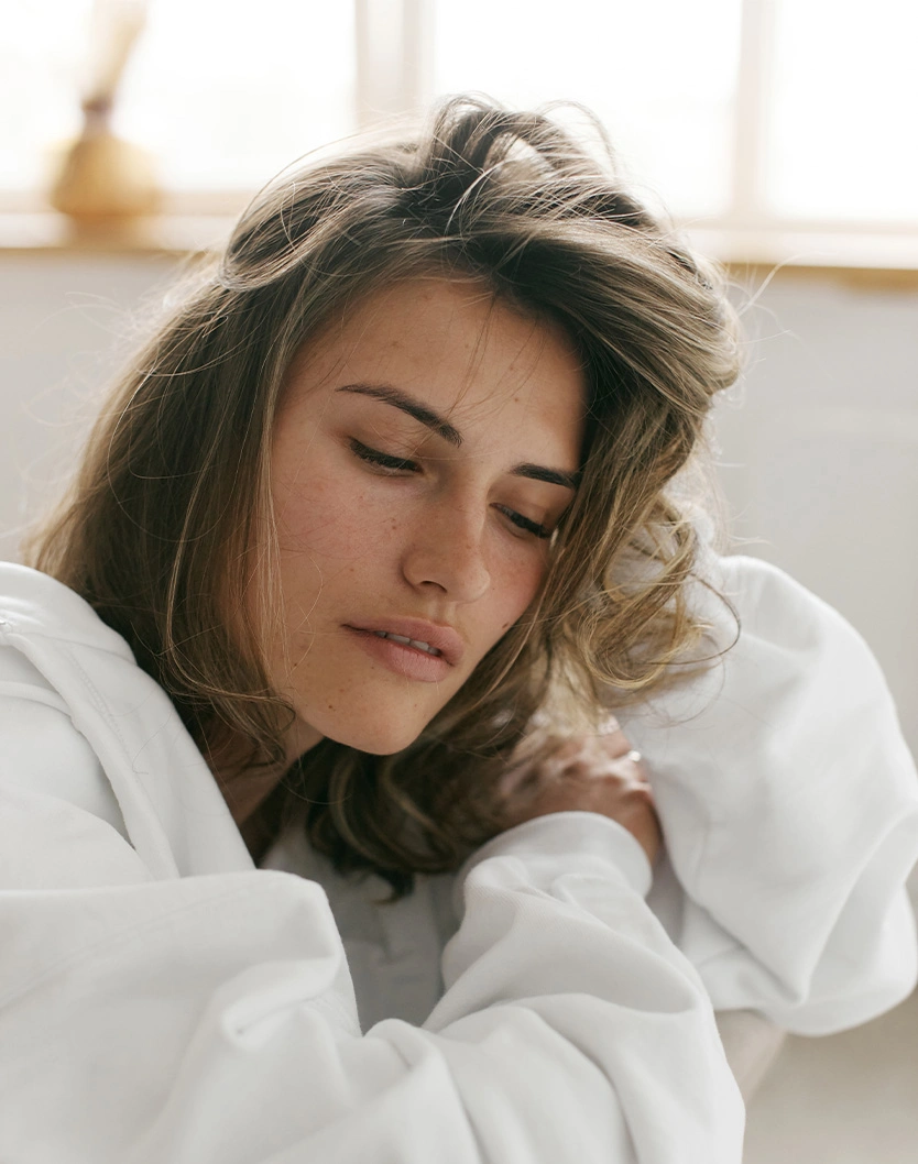 Woman with wavy brown hair relaxing in white bathrobe looking down peacefully in bright natural light