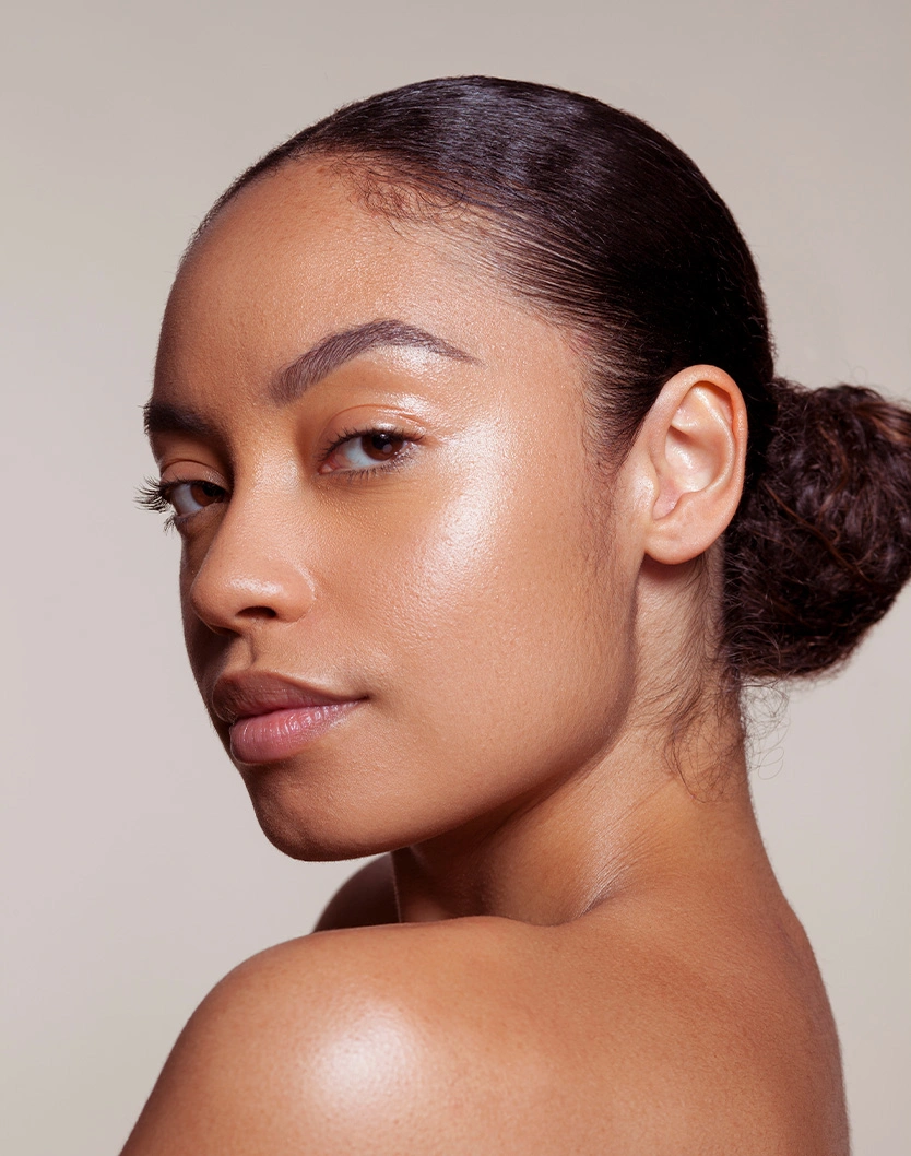 Woman with natural makeup and slicked back hair in bun showing glowing skin and bare shoulders against neutral background