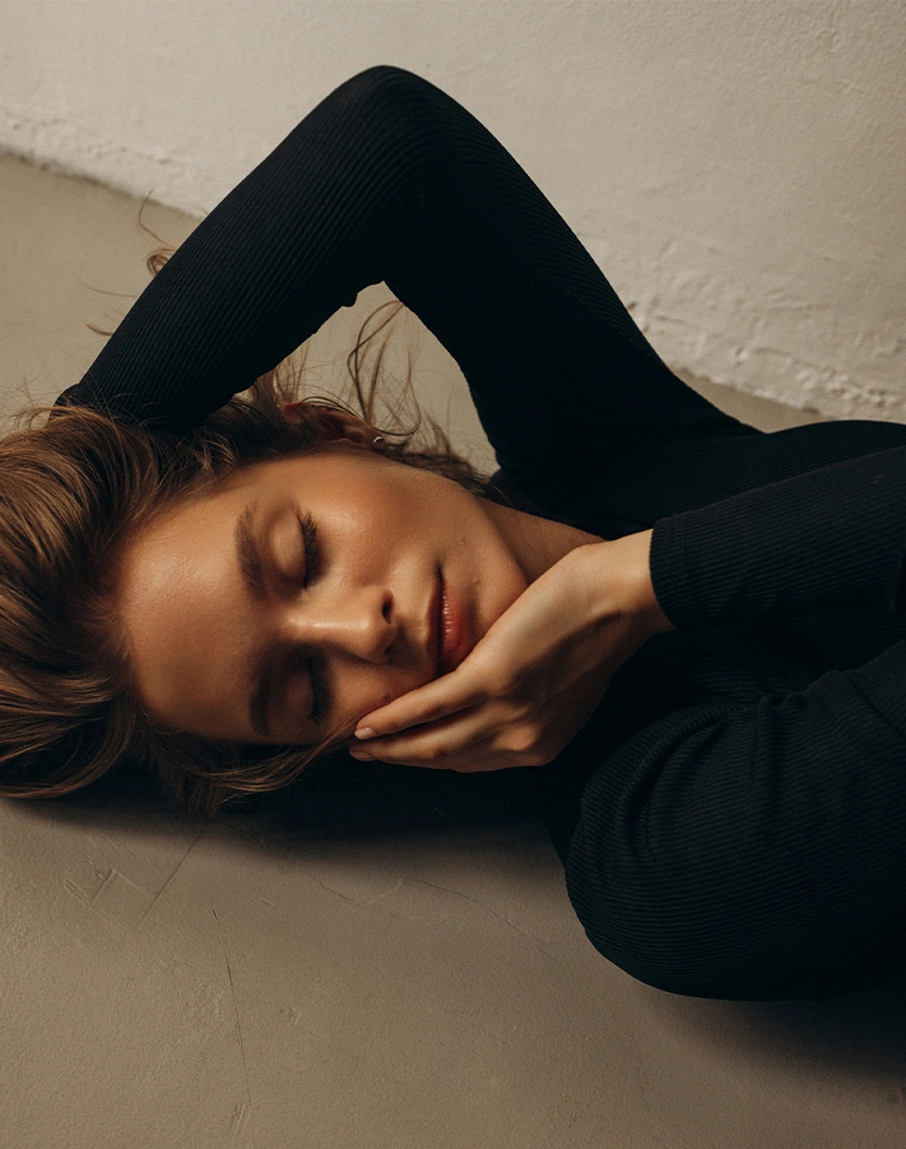 Woman with dark hair lying down in black clothing against neutral background in contemplative pose