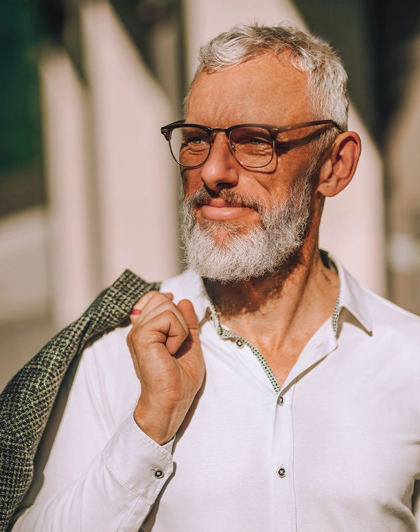 Distinguished mature man with gray beard and glasses smiling outdoors in white shirt holding green jacket