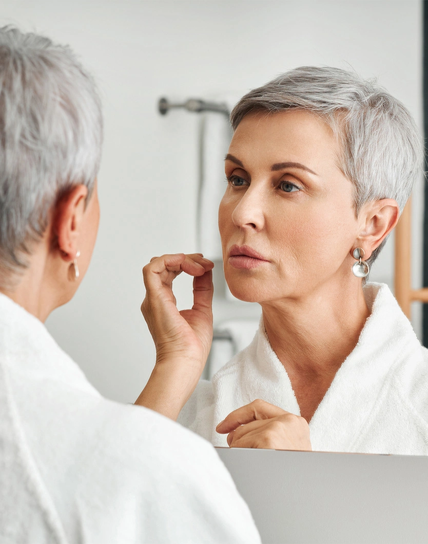 Woman with short gray hair examining her face in bathroom mirror while touching her skin