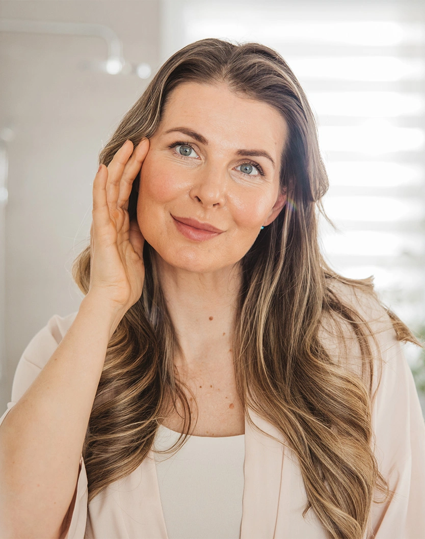 Woman with long brown hair touching face while smiling at camera in natural light setting