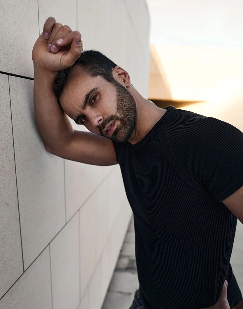 Man in black t-shirt posing against white tiled wall with arm raised above head in fashion portrait