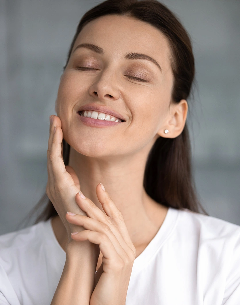 Woman with smooth skin touching face and smiling with eyes closed in white robe showing skincare results