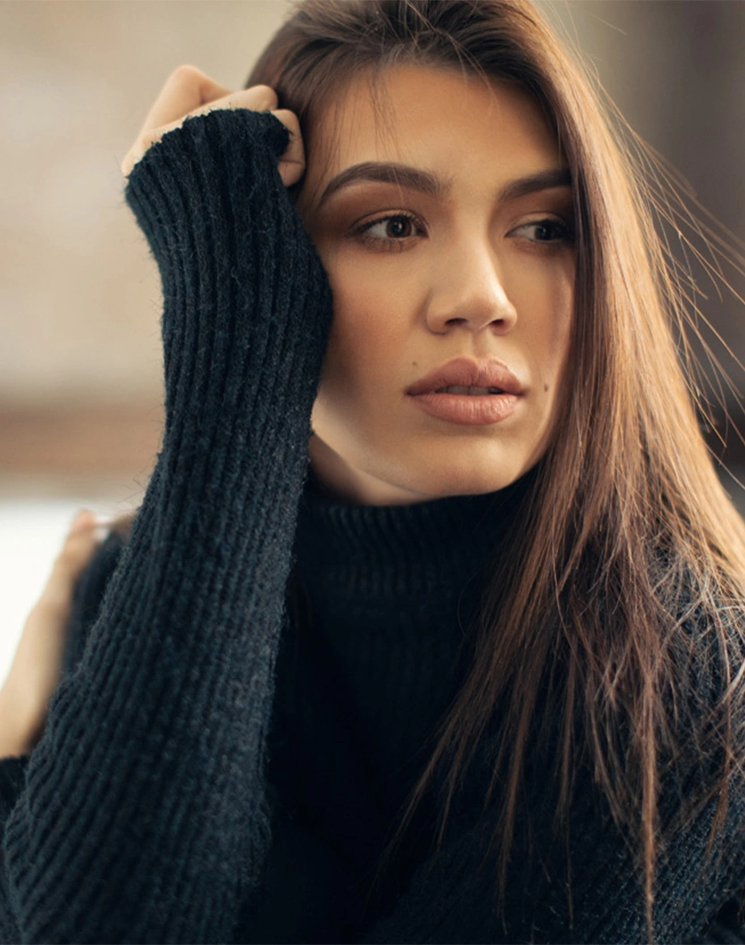 Woman with long brown hair wearing black turtleneck sweater posing with hand near face in natural lighting