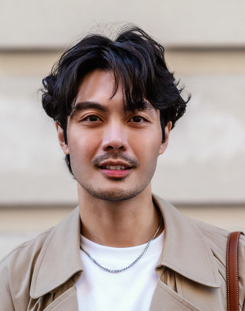 Young man with curly black hair and mustache wearing beige jacket and white shirt outdoors